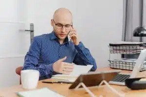 A fleet manager in a professional office environment, talking on a mobile phone while cross-referencing a driver qualification file and a digital monitoring dashboard on a tablet and laptop.
