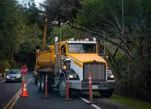 A yellow and white commercial dump truck stopped on the shoulder of a two-lane road surrounded by orange traffic cones, with an oncoming car passing a construction worker holding a stop sign in a wooded area.