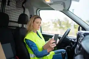 woman using tablet in van A female commercial fleet driver wearing a yellow safety vest inside a delivery van cab, using a mobile app on her smartphone to submit a real-time incident report.
