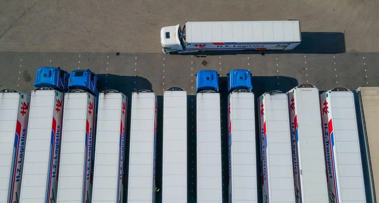 An aerial drone shot of commercial logistics trucks parked in a row, with one white semi-truck driving independently above them, representing unbundled fleet management solutions.