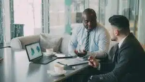 Two corporate risk management professionals discussing fleet cost reductions and insurance claims data on a laptop and charts over coffee.