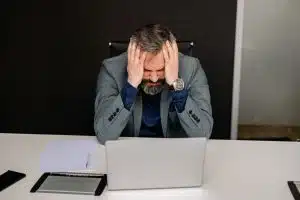 stressed businessman staring at laptop A frustrated fleet safety manager sitting at a desk with his head in his hands, looking stressed while working on a laptop with manual incident reports.
