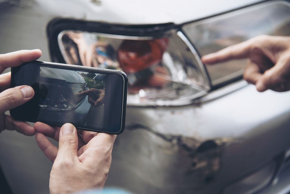 Close-up of a person taking a photo of a damaged car headlight and bumper with a smartphone.