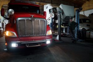 A mechanic standing next to a large red semi-truck parked inside a commercial repair garage.